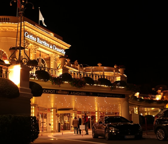 A luxurious casino at night, illuminated with warm, golden lights. The building features elegant architecture with columns and decorative elements. A few people are walking towards the entrance, and there are a couple of parked cars nearby.